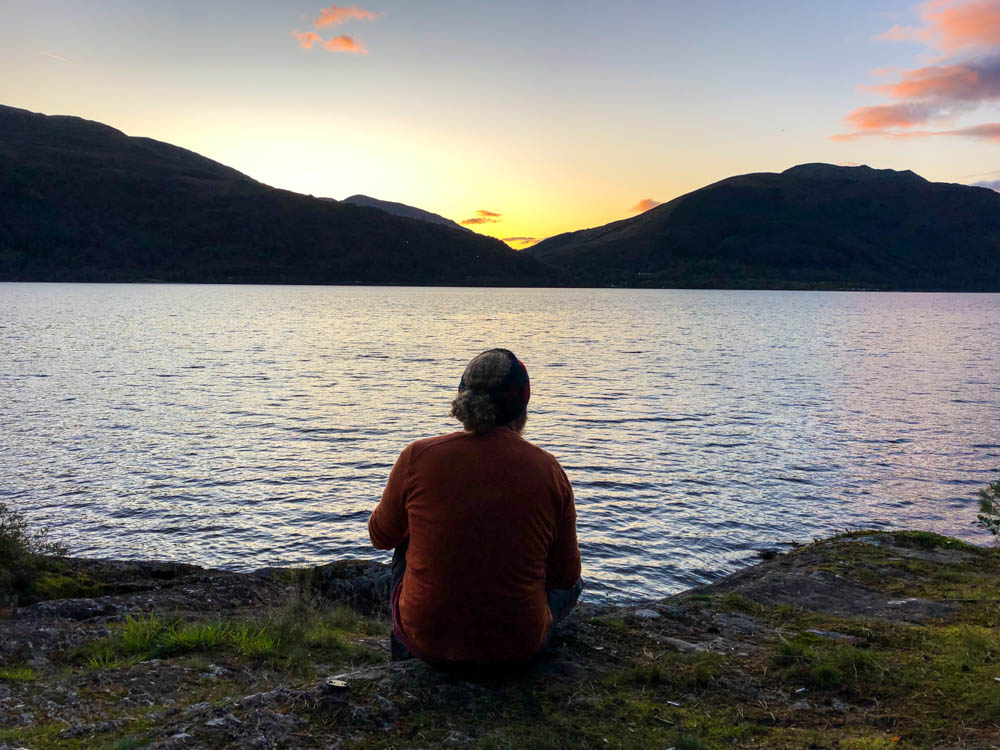 Julian sitzt auf dem Boden, vor ihm ist ein See, er blickt auf das gegenüberliegende Gebirge. Die Sonne ist gerade untergegangen und alles leuchtet leicht orange. Trekking West Highland Way Schottland