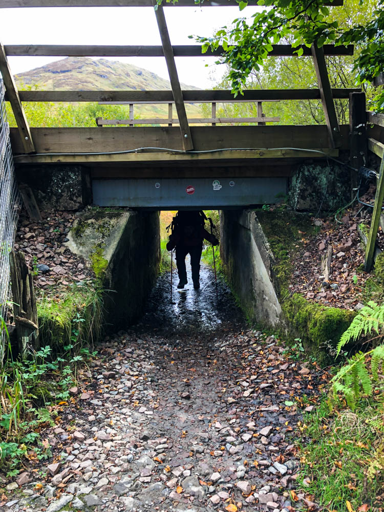 Julian wandert durch eine Unterführung, welche jedoch nur halb so ist wie er selbst. Er muss gebückt durch laufen. Trekking West Highland Way