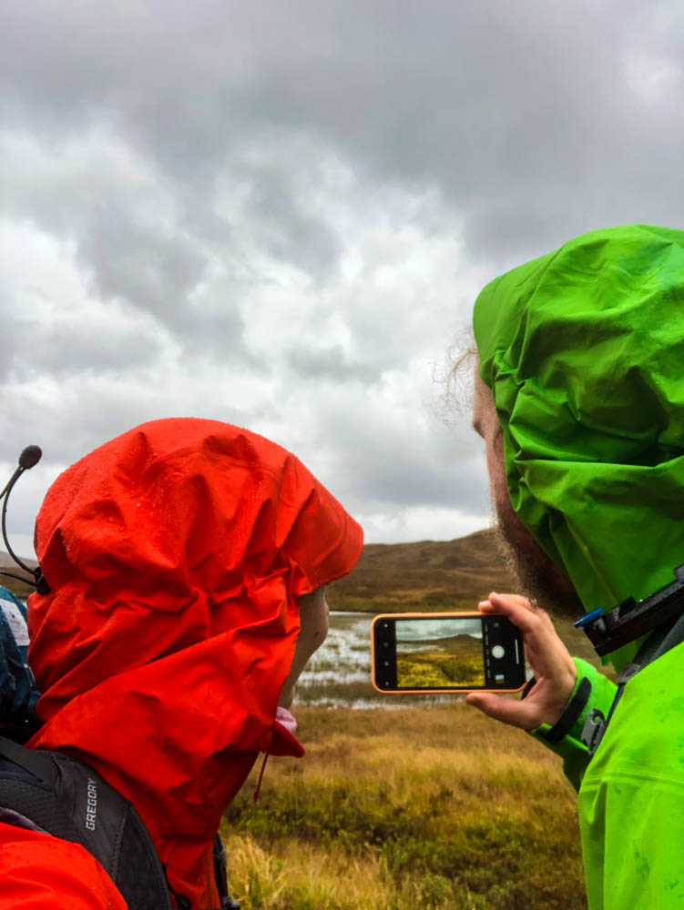 Aufnahme von Melanie und Julian wie sie mit ihrem Handy ein Foto der Landschaft machen. Der Himmel ist bewölkt und es regnet. West Highland Way Schottland