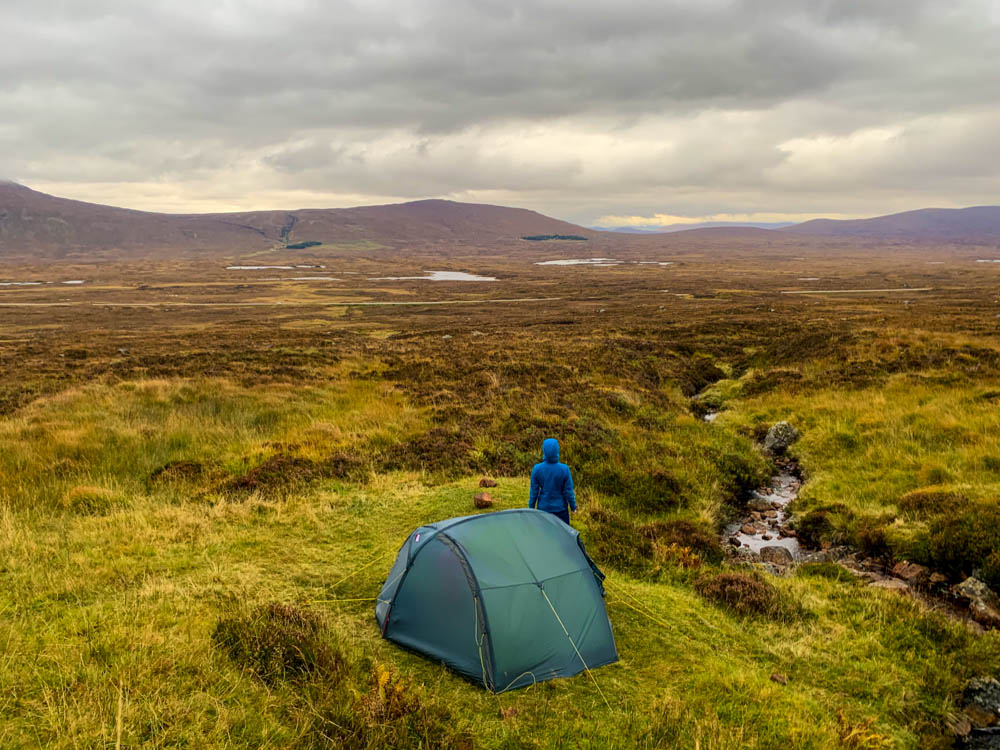 Melanie steht neben dem aufgebauten Zelt mitten in den Highlands und blickt in die Ferne. Trekking West Highland Way Schottland