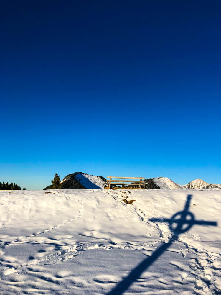 Das Gipfelkreuz des Wank wirft gegenüber im Schnee einen Schatten. Der Himmel ist kräftig blau - Winter in den Alpen