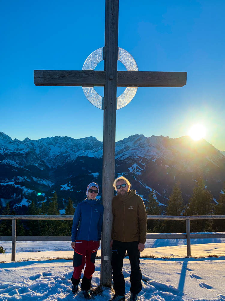 Melanie und Julian stehen am Gipfelkreuz vom Wank in Winterlandschaft. Der Himmel ist kräftig blau und die Sonne ist mit im Bild