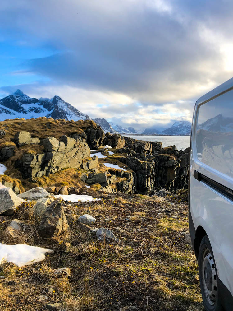 Van Vivaldi steht an einem Fjord auf den Lofoten im April. Es ist leicht bewölkt und die Sonne scheint. Die Berge spiegeln sich auf dem Van.