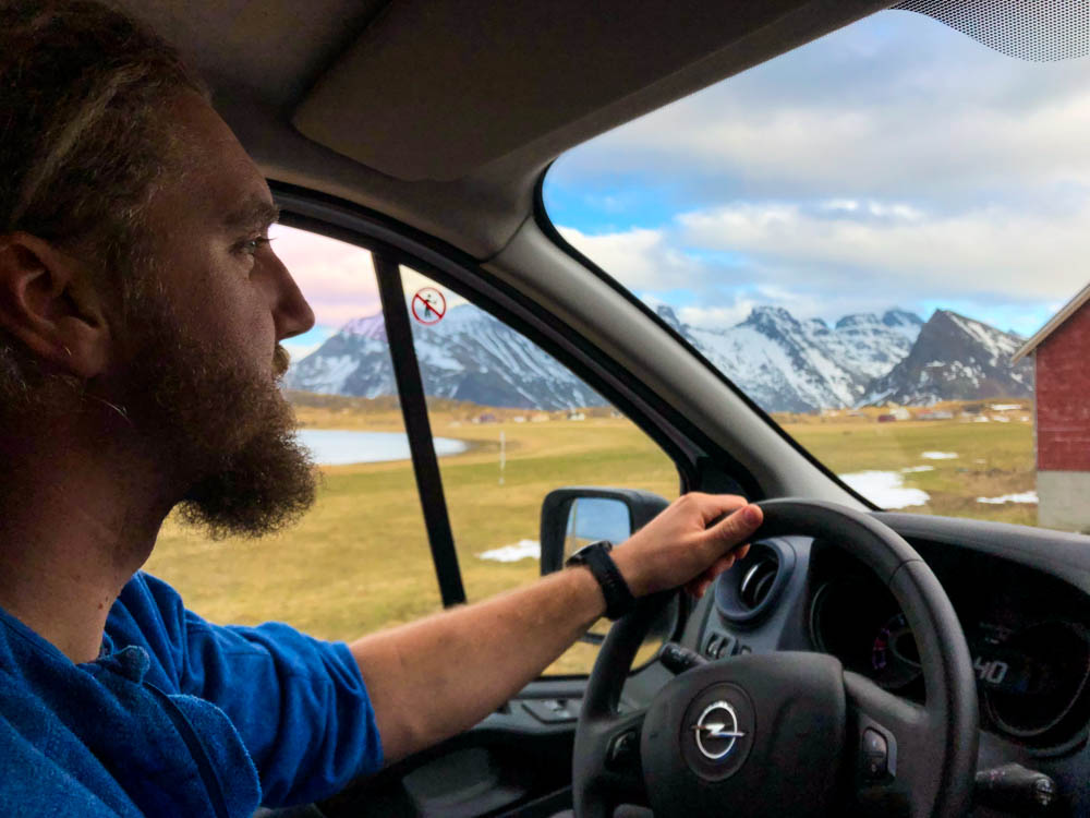 Julian sitzt am Steuer von Van Vivaldi und Fährt durch die schöne Landschaft der Lofoten. Es sind ein paar schneebedeckte Berge zu sehen sowie das satte grün der Wiesen.