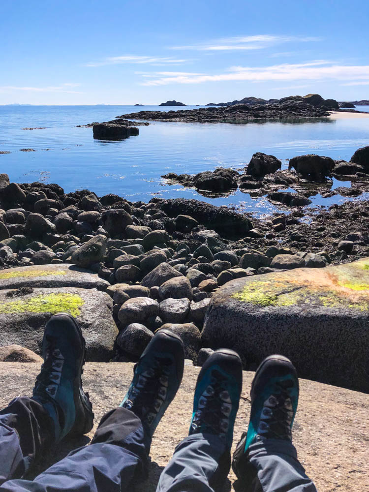Es sind die Füße mit Wanderstiefeln von Melanie und Julian zu sehen. Sie halten diese in Richtung des Atlantiks auf den Lofoten, welcher direkt vor ihnen liegt.