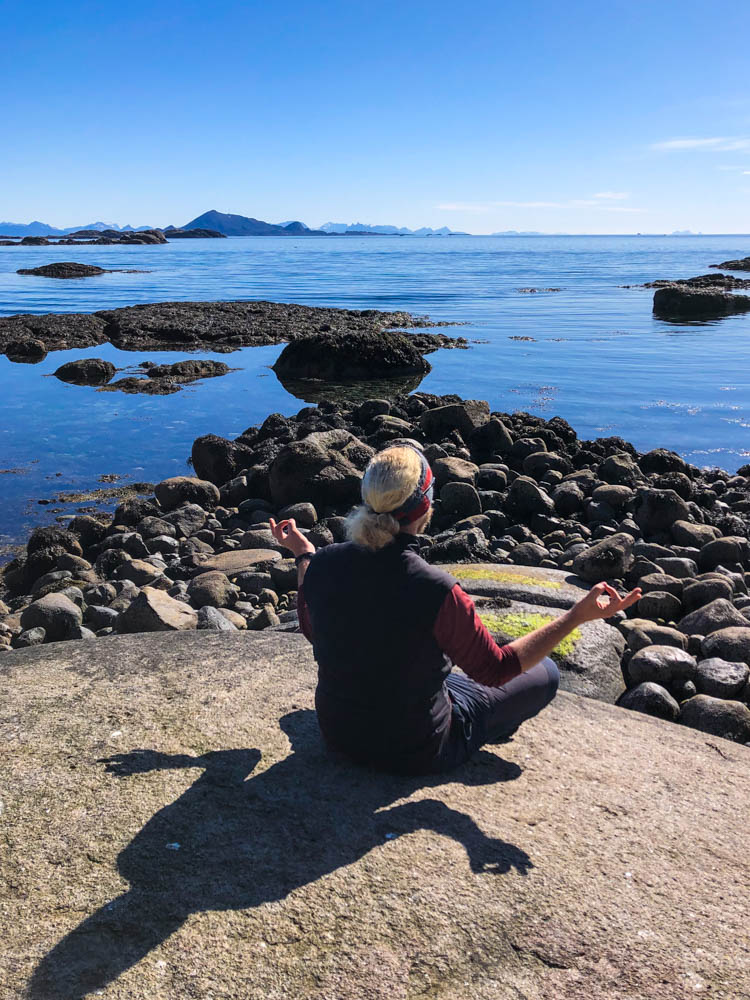 Julian beim meditieren am Atlantik auf den Lofoten. Der Himmel strahlt kräftig blau genau wie das Wasser des Meeres.