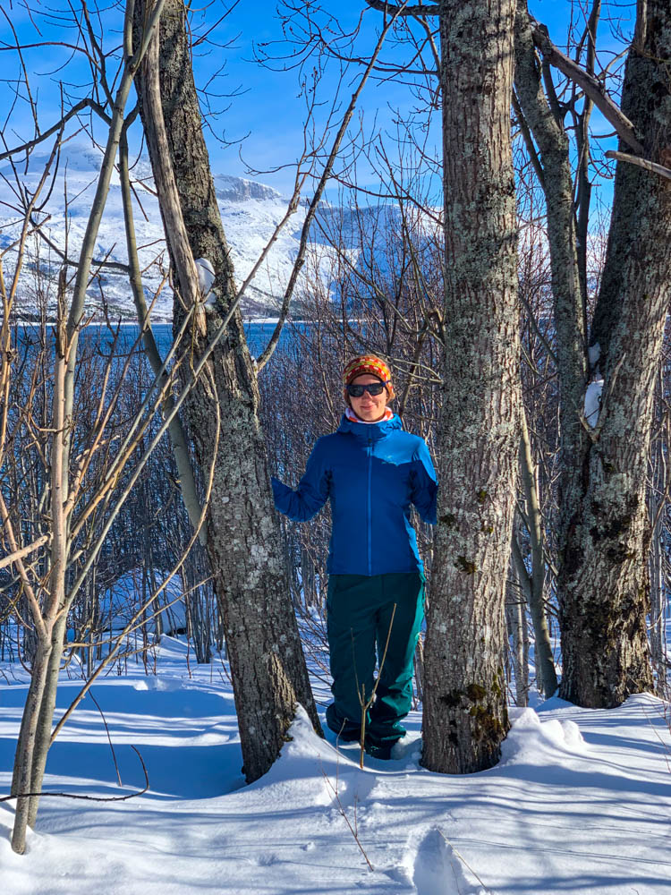 Melanie steht zwischen zwei Bäumen im Schnee und grinst in die Kamera. Im Hintergrund ist der Atlantik zu sehen sowie verschneite Berge. Der Himmel ist kräftig blau Anfang April in Nordnorwegen