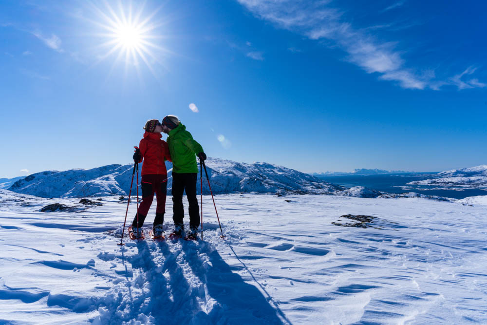 Schneeschuhwanderung Nordnorwegen Anfang April. Melanie und Julian küssen sich, im Hintergrund verschneite Berglandschaft sowie Sonnenstern, der Himmel ist kräftig blau.