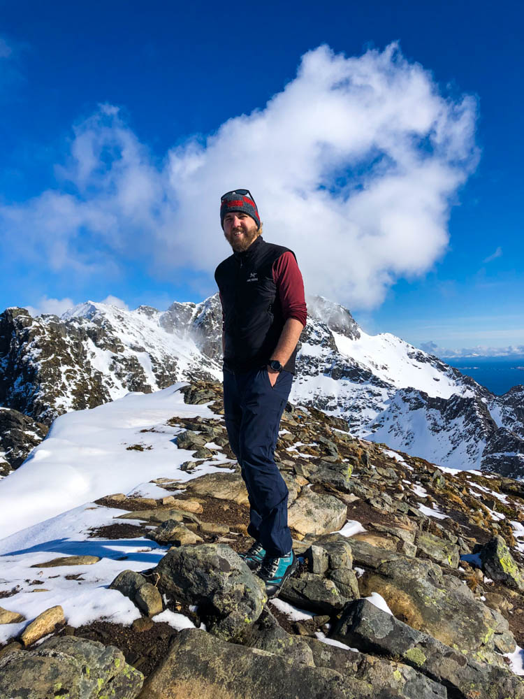 Julian am Gipfel des Festvågtinden bei Henningsvaer im April. Der Himmel ist kräftig blau und es liegt Schnee auf den Bergen. Julian grinst in die Kamera