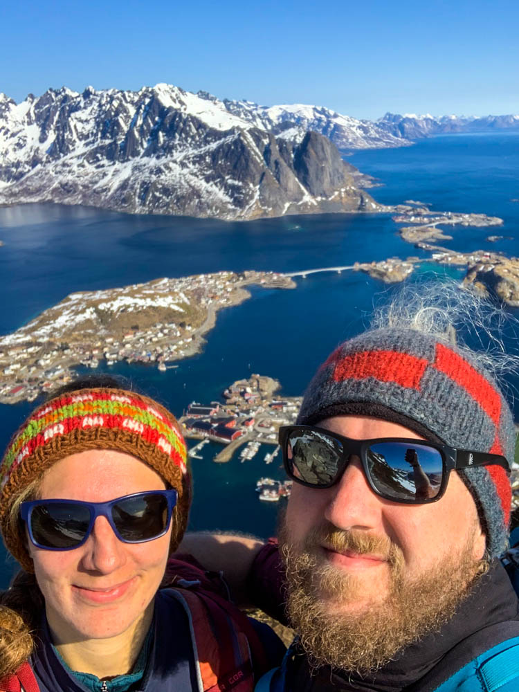 Selfie von Melanie und Julian Reinebringen Lofoten im April. Im Hintergrund ist Reine zu sehen, der Himmel ist strahlend blau genau wie der Reinefjord und der Atlantik.