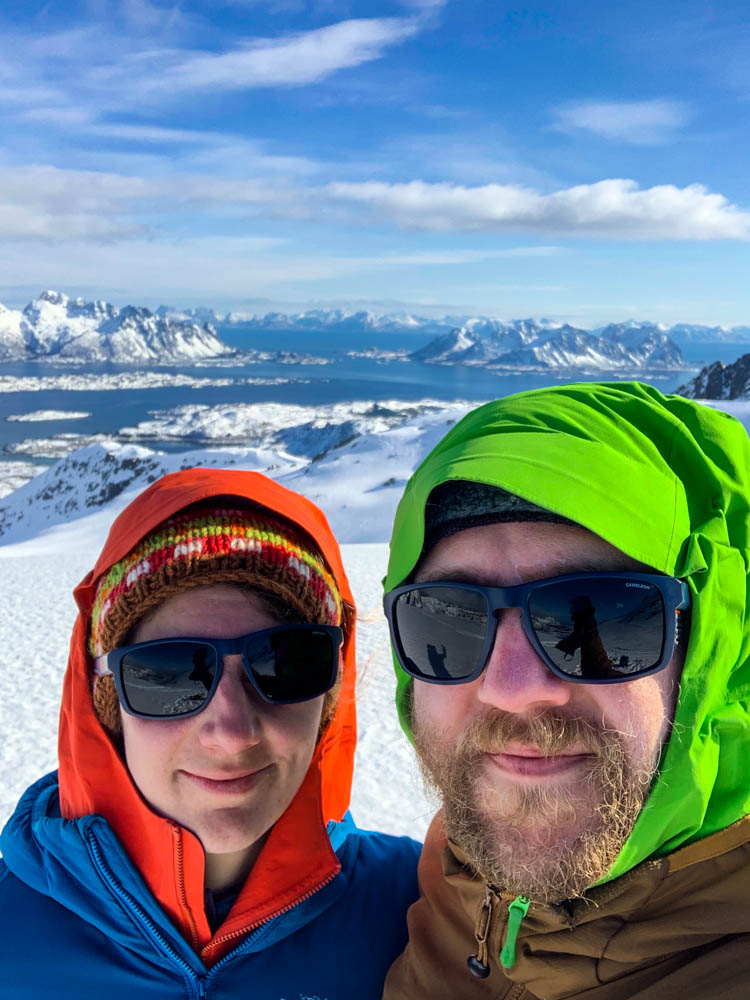 Selfie von Melanie und Julian bei einer Splitboardtour am Rundfjellet auf den Lofoten im April. Im Hintergrund ist ein schönes Bergpanorama zu sehen sowie eine Fjord Landschaft. Der Himmel ist nur leicht bewölkt.
