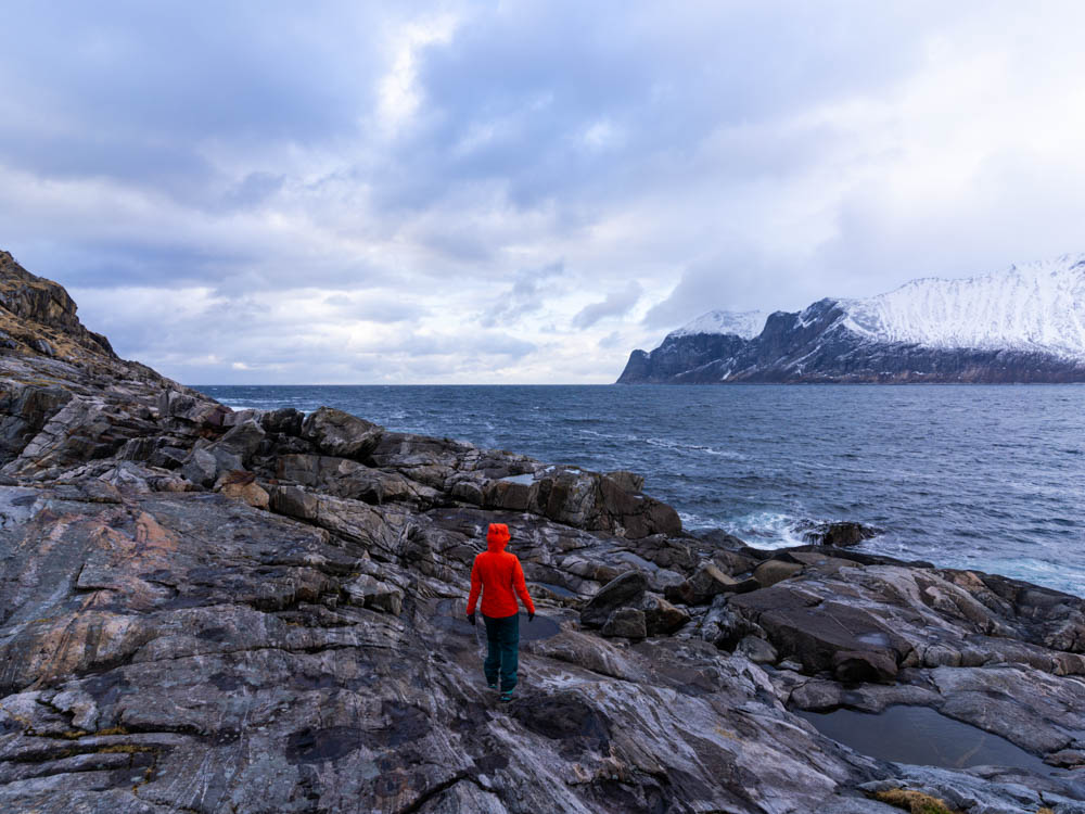 Melanie steht auf Steinen an der Küste und blickt auf den Atlantik sowie die gegenüberliegende verschneite Berglandschaft.