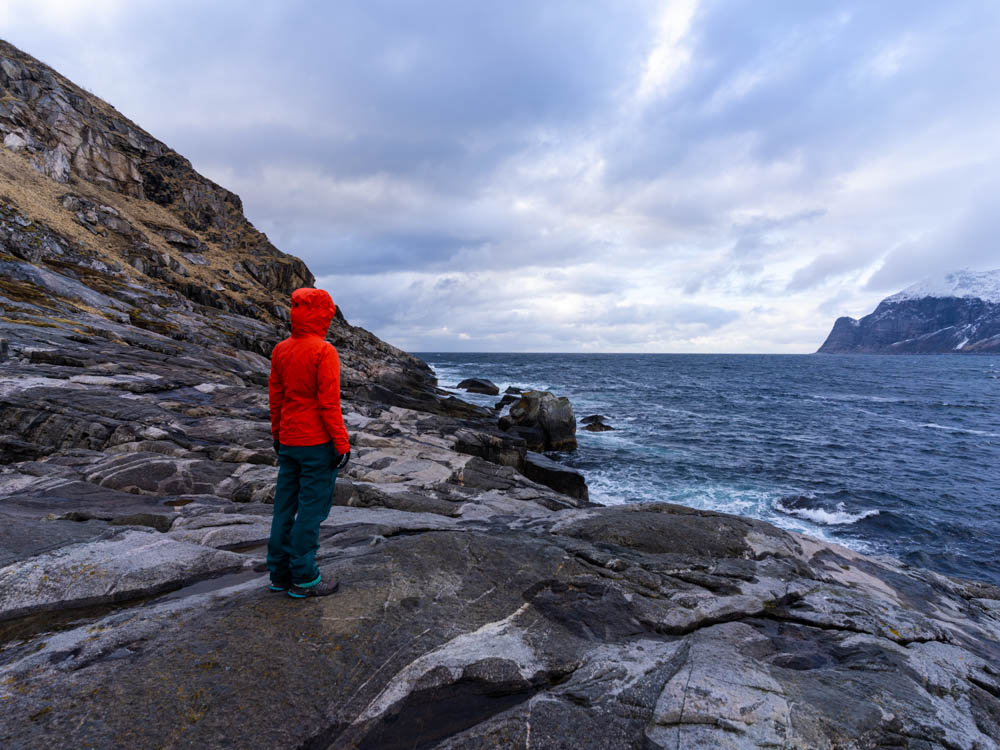 Melanie steht auf Steinen an der Küste und blickt auf den Atlantik sowie die gegenüberliegende verschneite Berglandschaft.
