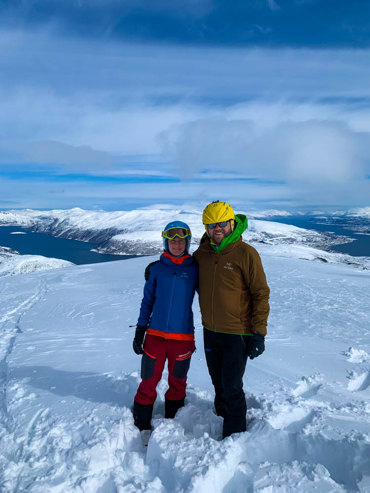 Melanie und Julian stehen auf dem Gipfel vom Steinskarstinden. Hinter ihnen ist eine verschneite Berglandschaft zu sehen.