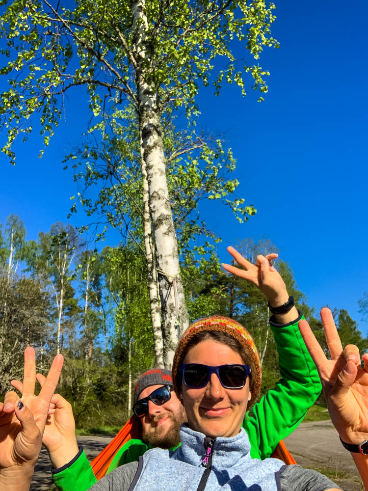 Melanie und Julian liegen in Schweden gemeinsam in einer Hängematte und zeigen mit ihren Händen das Peace-Zeichen. Es ist ein Wald hinter ihnen zu sehen sowie der strahlend blaue Himmel