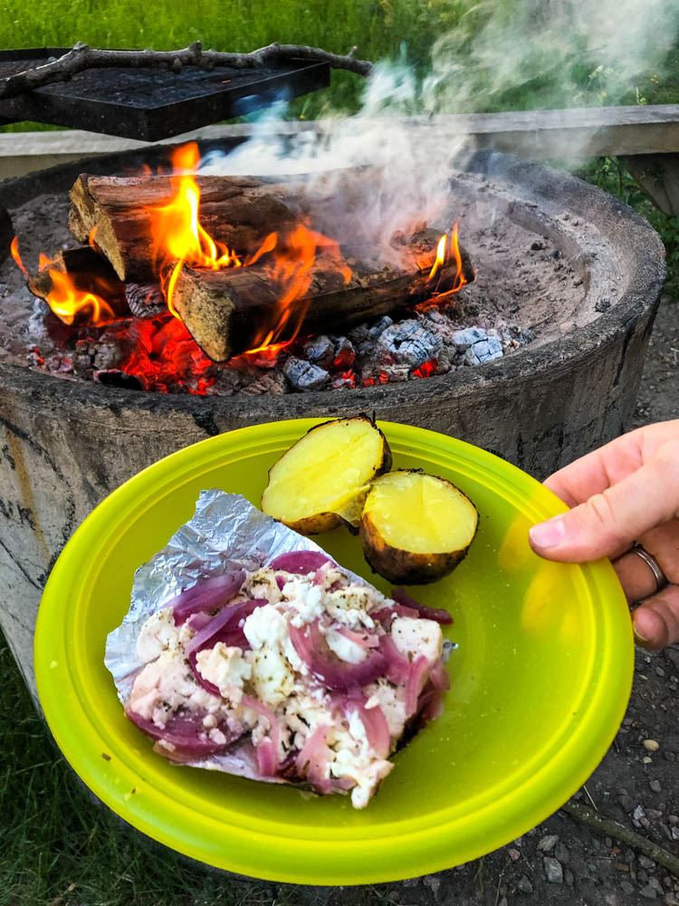Es wird das Essen des Abends vom Lagerfeuer präsentiert. Auf einem Teller sind Feta, Zwiebeln und Kartoffeln zu sehen. Das Feuer brennt im Hintergrund