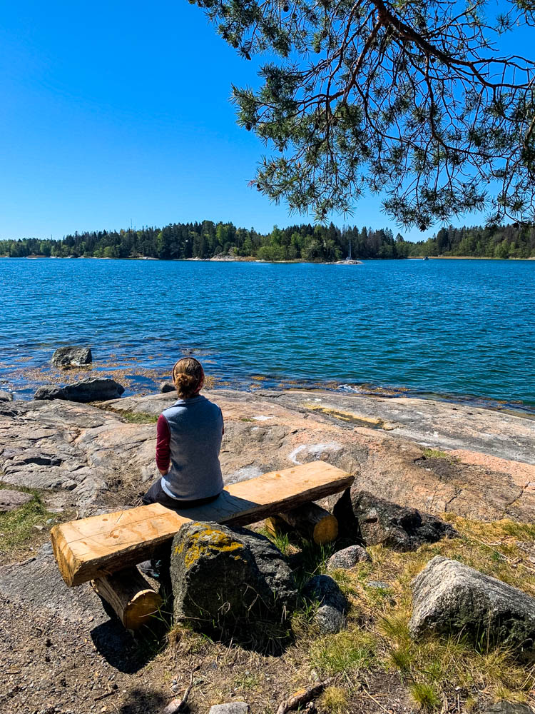 Melanie sitzt auf einer Bank in Schweden und blickt auf die Ostsee. Der Himmel ist kräftig blau.