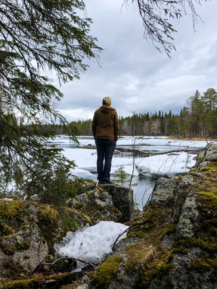 Julian steht auf einem kleinen Felsen und blickt in die Natur vor ihm. Auf einem Fluss liegt noch Schnee, rund herum ist ein Wald. Frühling in Nordschweden