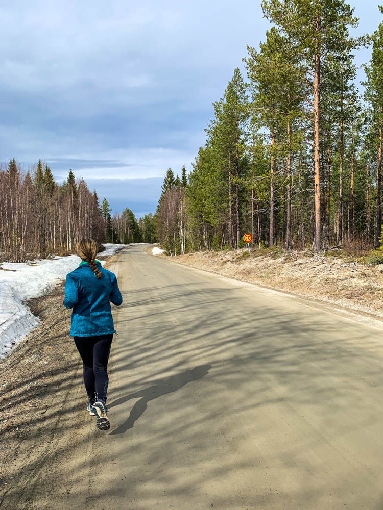 Melanie beim Joggen in Nordschweden. Sie läuft auf einer Straße, rund herum ist ein Wald zu sehen und ein paar Schneereste. Frühling in Nordschweden.