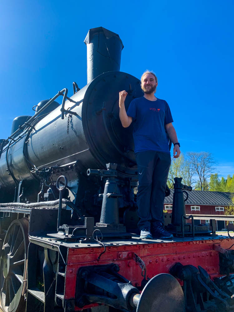 Julian steht auf einer Lokomotive in Nordschweden im Frühling und hebt den Arm. Der Himmel ist kräftig blau