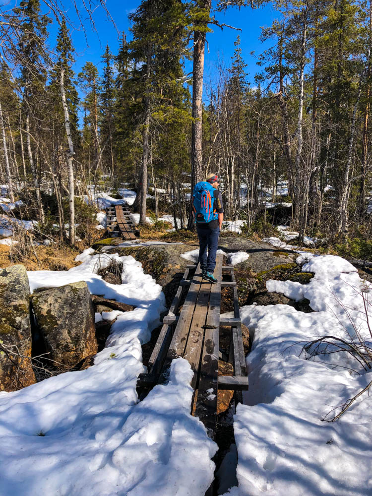 Julian läuft über eine Brücke über einen Fluss im Moor. Es liegt überall Schnee um ihn herum im Wald. Muddus Nationalpark in Nordschweden im Frühling