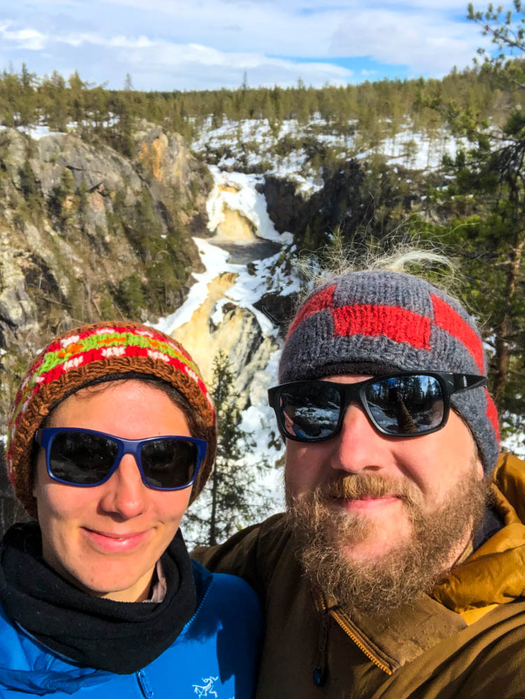 Selfie von Melanie und Julian im Muddus Nationalpark in Nordschweden. Hinter ihnen ist ein großer Wasserfall zu sehen. Es ist Frühling, zwischen dem Wald sind noch einige Schneefelder zu sehen.