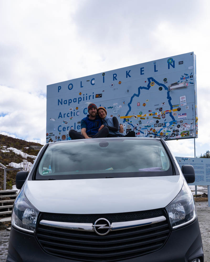 Van Vivaldi parkt unter dem großen Schild des Polarkreises in Nordschweden. Melanie und Julian sitzen auf dem Dach des Vans und halten sich im Arm.