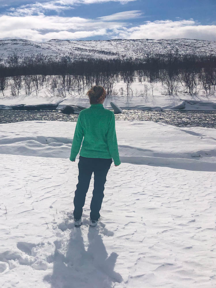 Melanie steht im Schnee in Nordskandinavien und blickt auf die weiße Landschaft. Vor ihr ist ein weißer, vereister Fluss.