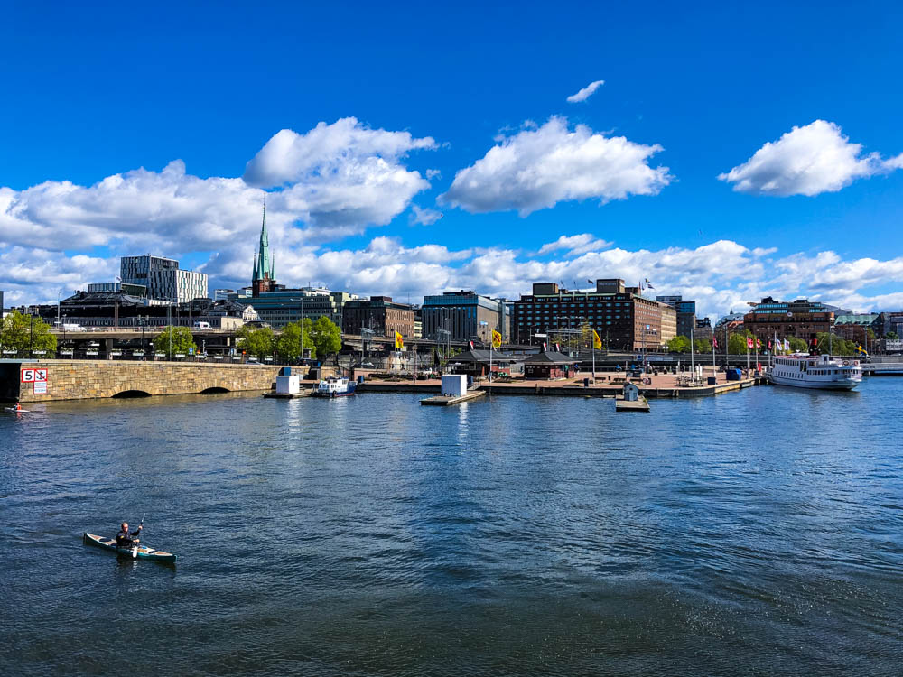 Blick auf einen Fluss in Stockholm sowie die dahinterliegende Stadtfront. Der Himmel ist kräftig blau und es sind ein paar Wolken zu sehen.