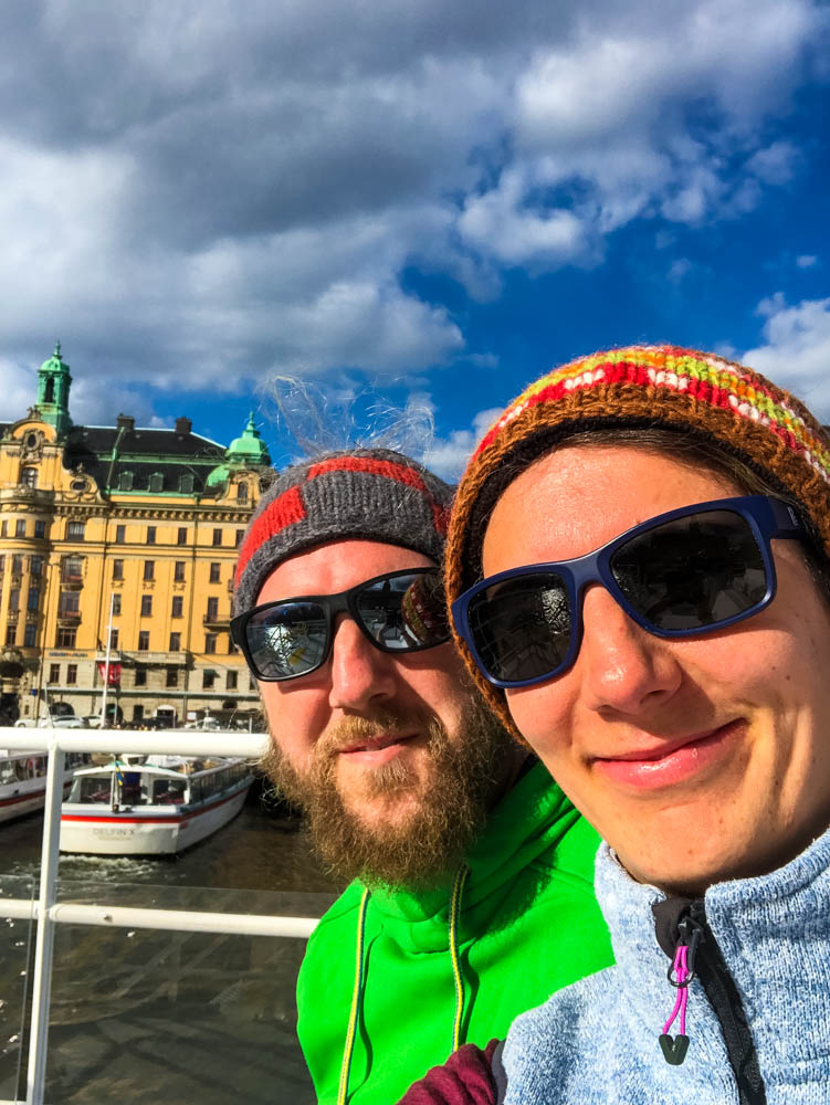 Selfie von Melanie und Julian auf der Fähre in Stockholm. Sie grinsen beide in die Kamera, hinter ihnen sind anliegende Boote sowie ein Gebäude zu sehen.