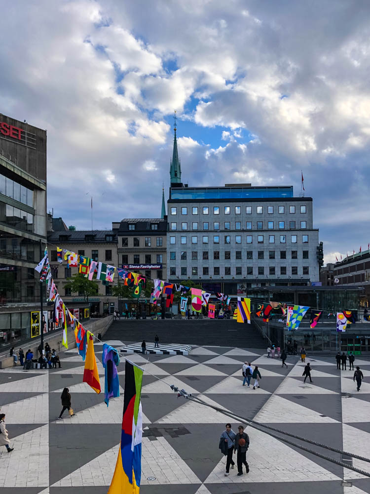 Reges Treiben auf einem Platz in Stockholm. Es hängen viele verschiedene Flaggen an mehreren Seilen über diesem Platz.