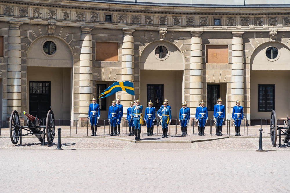 Wachwechsel im Hof des Königspalastes in Stockholm. Wachleute stehen aufgereiht, vor ihnen steht einer der Wachleute mit einer schwedischen Fahne in der Hand