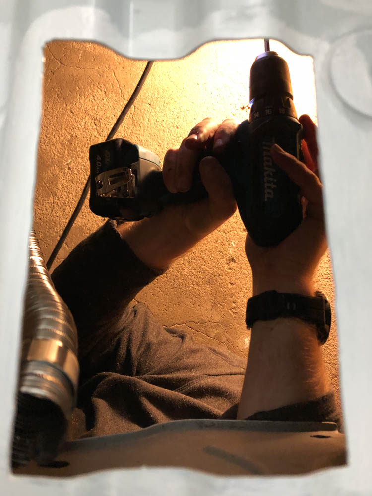 Julian liegt unter dem Auto und bohrt ein Loch für die Halterung vom Auspuff. Die Aufnahme wurde durch den Bodenauschnitt im Van gemacht welcher für die Standheizung eingesägt wurde.