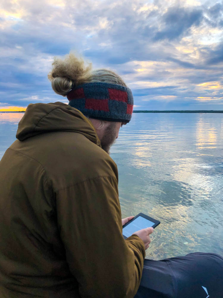 Julian sitzt direkt an der Ostsee in Südschweden und liest ein Buch auf seinem E-Book. Im Hintergrund geht die Sonne unter.
