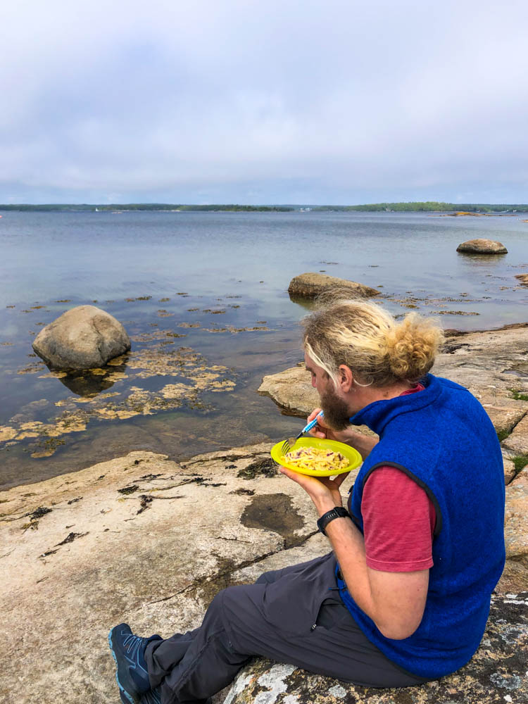 Julian sitzt auf einem Stein direkt an der Ostsee und isst Kässpätzle aus einem Teller.