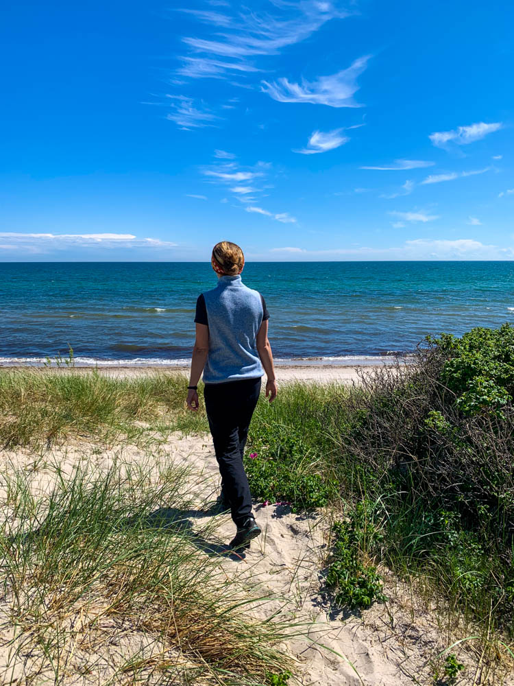 Melanie läuft im Sand der Ostsee hingegen. Im Sand sind ein paar grüne Pflanzen zu sehen, der Himmel ist kräftig blau. Ostsee bei Ystad Südschweden