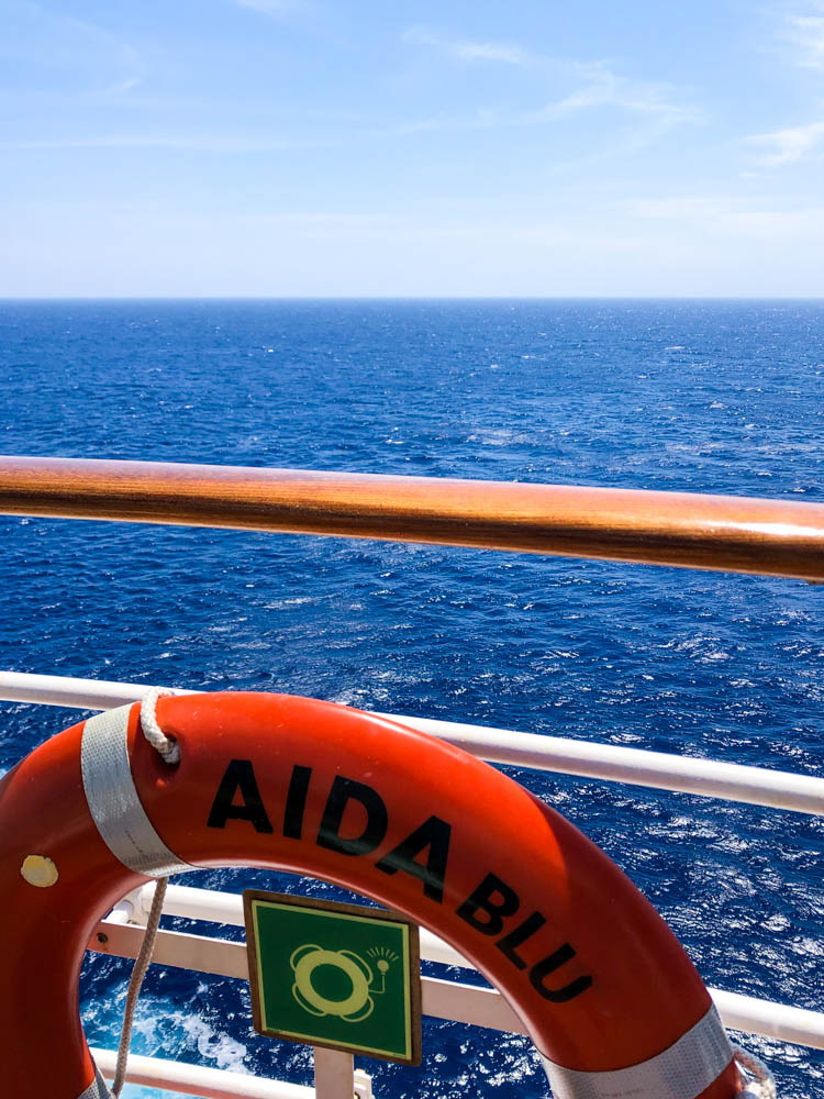 Ein Rettungsring auf welchem AIDAblu steht hängt an der Reling. Dahinter ist der weite Ozean zu sehen.