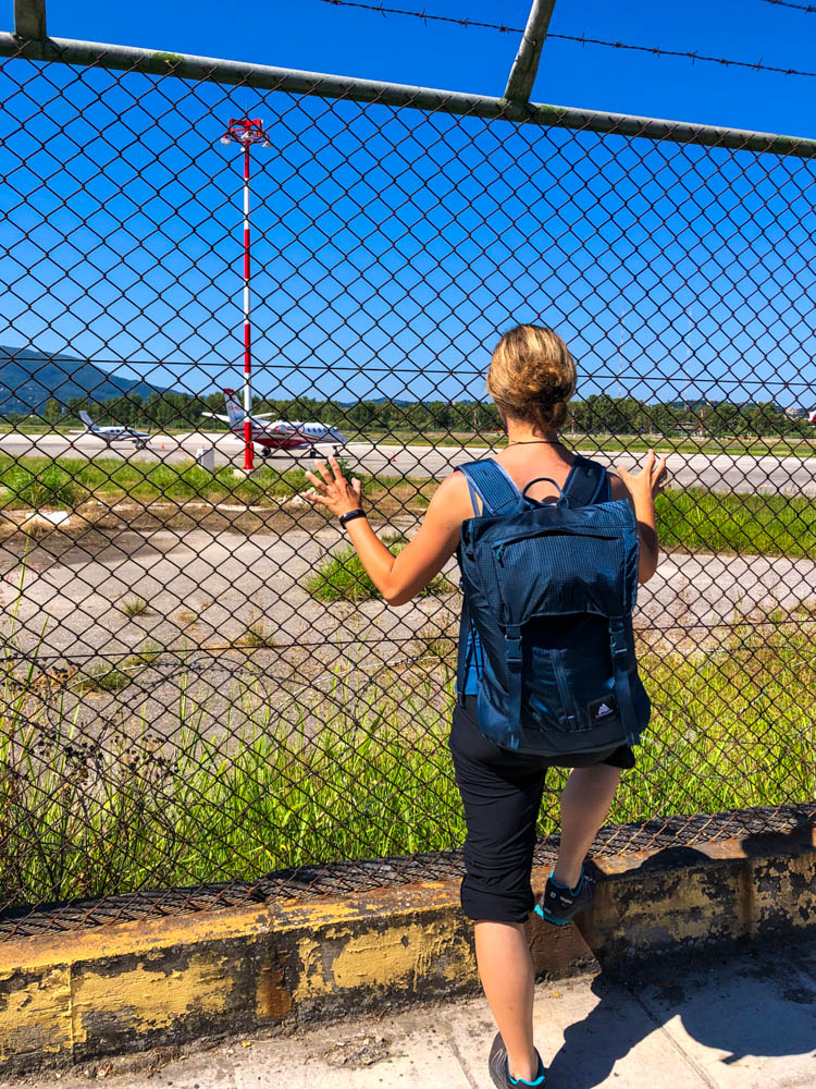 Melanie steht am Zaun vom Flughafen in Korfu und hält sich am Zaun fest. Es sieht so aus als würde sie wo ausbrechen wollen