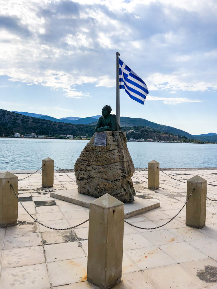 Eine Statue auf einem Fels. Dahinter weht die Griechische Nationalfahne.