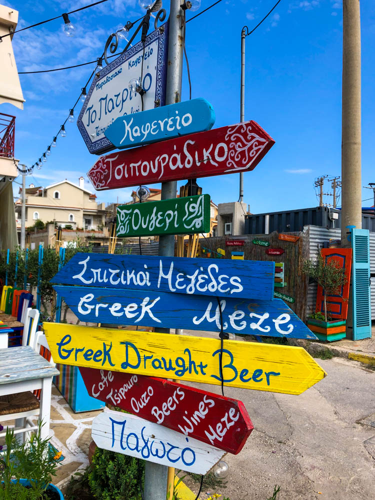 Ein bunter Wegweiser aus Holz der in viele verschiedenen Richtungen zeigt. Es für uns kaum etwas lesbar, da es in griechischer Schrift geschrieben ist. Nur das Greek Draught Beer kann gut gelesen werden .
