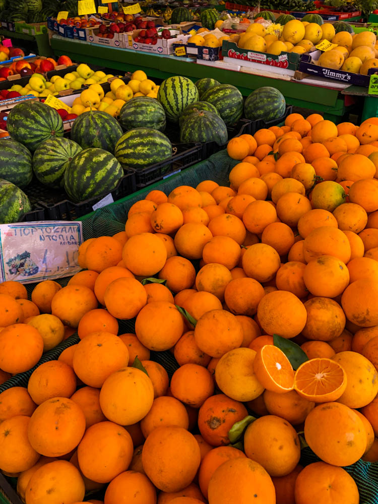 Ein Obstatnd mittden in der Stadt. Es sind Zitronen, Melonen und Orangen zu sehen. Im Hintergrund noch ein paar andere Obstsorten. Das Bild ist knallbunt.