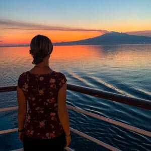 Melanie blickt auf den Ätna in Sizilien, hinter welchem die Sonne untergegangen ist. Sie steht auf einem Schiff im Mittelmeer und die rote Farben des Himmels spiegeln sich im Wasser. Horizont erweitern