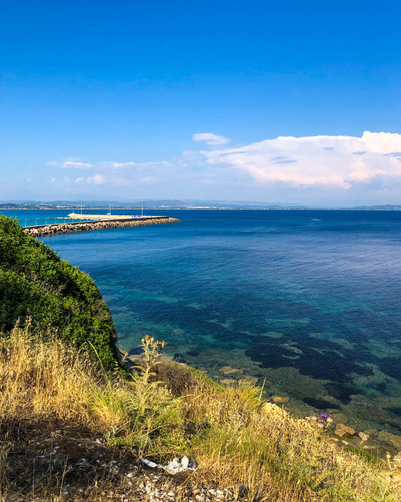 Aufnahem vom Küstenabschnitt und einem Stück der Hafenmauer in Katakolo. Das Bild ist von einer Anhöhe gemahct. Das Wasser spiegelt herrlich blau und das Grün der Pflanzen ist sehr kräftig. Ein schöner Kontrast. Hier kann man super seinen Horizont erweitern zumindest Wortwörtlich vom Ausblick her.
