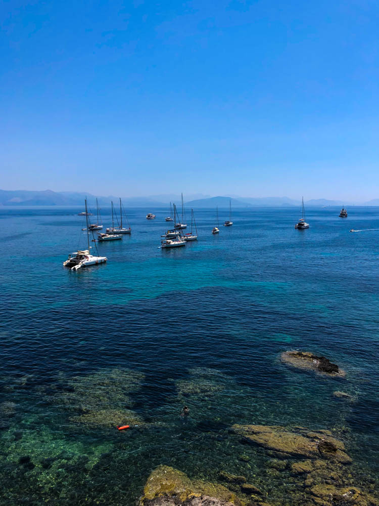 Küste in Korfu. Das Wasser schimmert in verschiedenen Blautönen und es liegen ein paar Schiffe darauf. Der Himmel ist kräftig blau.