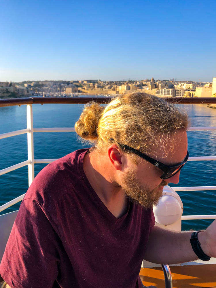 Julian sitzt am Tisch und genießt dein Frühstück während das Schiff im Hafen von Valetta anlegt. im Hintergrund ist die Stadt zu sehen.