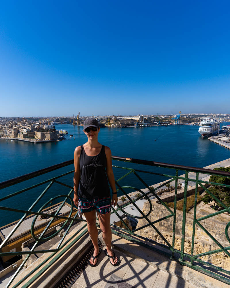 Melanie steht am Geländer auf einer Aussichtsplattform der Altstadt in Valetta und lächelt in die Kamera. Im Hintergrund ist die Stadt und der Hafen, mit der AIDAblu zu sehen. Ein Blick, der den Horizont erweitert.