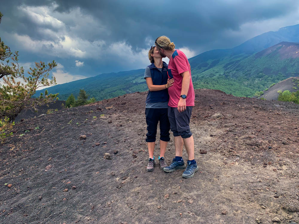 Melanie und Julian stehen an einem kleinen Hügel und küssen sich. Im Hintergrund ist der wolkenverhangene Ätna zu erahnen.