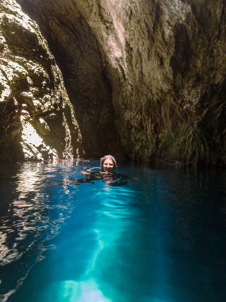 Melanie ist beim Tauchen in einer Grotte bei Paleokastritsa auf Korfu aufgetaucht. Sie zeigt das ok Zeichen und schaut glücklich in die Kamera. Das Wasser ist türkisfarben, die Höhle rund herum schimmert von der Sonne, die oben durch ein Loch hereinfällt.