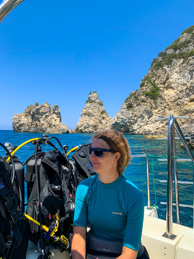 Melanie beim Tauchen bei Paleokastritsa auf Korfu. Sie sitzt auf einem Tauchboot in ihrem Neoprenanzug und schaut glücklich in die Ferne. Hinter steht Tauchequipment