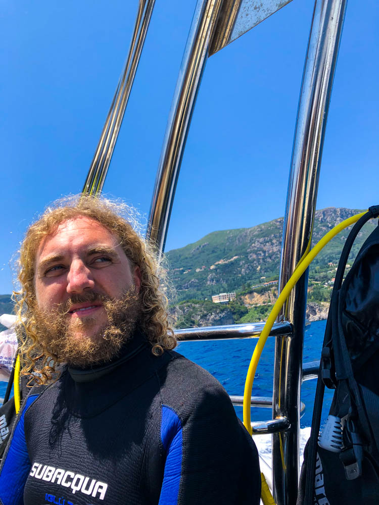 Julian beim Tauchen bei Paleokastritsa auf Korfu. Er sitzt auf einem Tauchboot in seinem Neoprenanzug und schaut in die Ferne. Der Himmel ist kräftig blau.